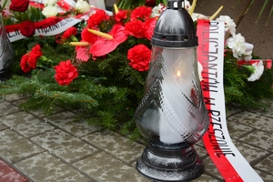 Płonący znicz, w tle wiązanki kwiatów.